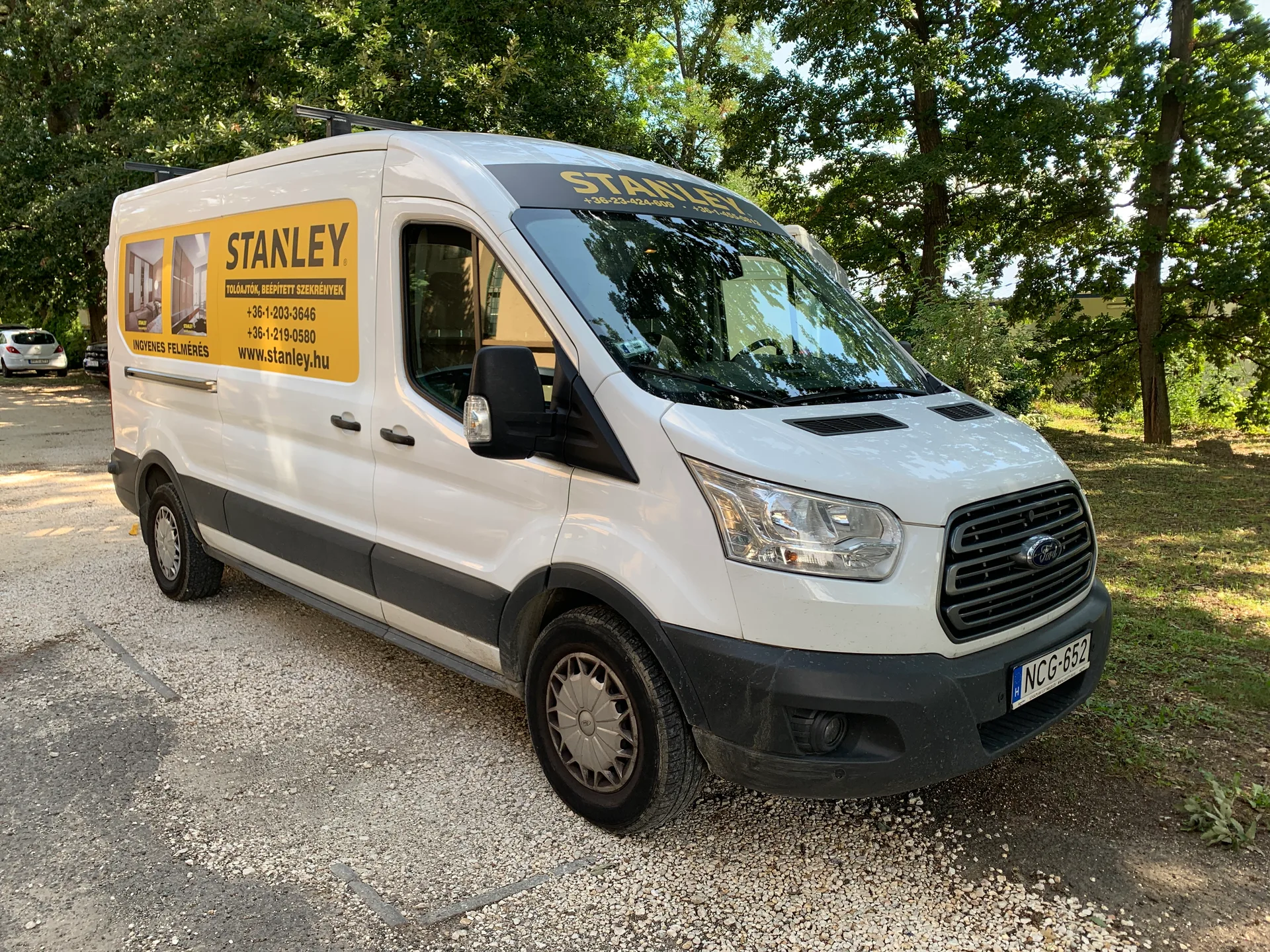 Fehér Stanley Ford Transit furgon háromnegyedes elülső jobb oldali nézete sárga-fekete vakablak-dekorral és szélvédő feletti Stanley felirattal